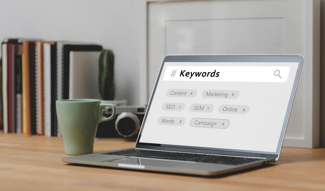 A laptop displaying a search for keywords related to SEO and marketing, placed on a wooden desk with a green coffee mug, books, and decor in the background.